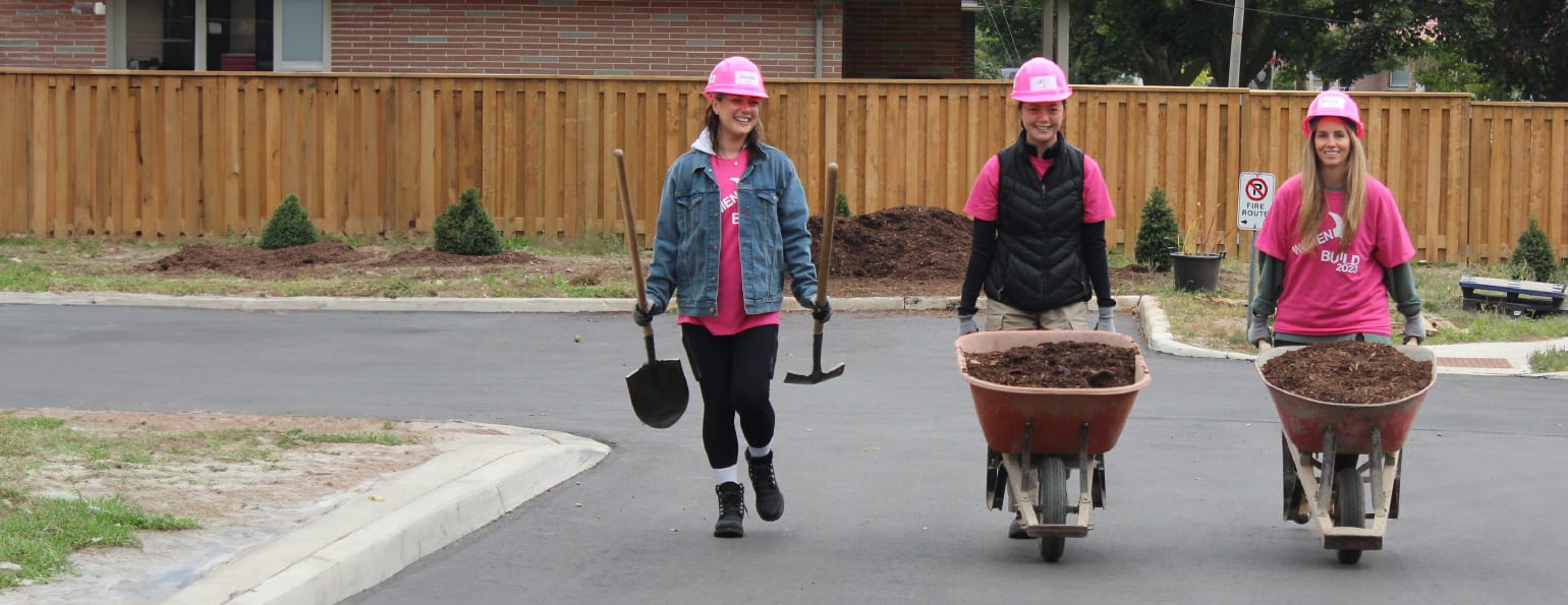 Three women walking with tools and wheel barrows